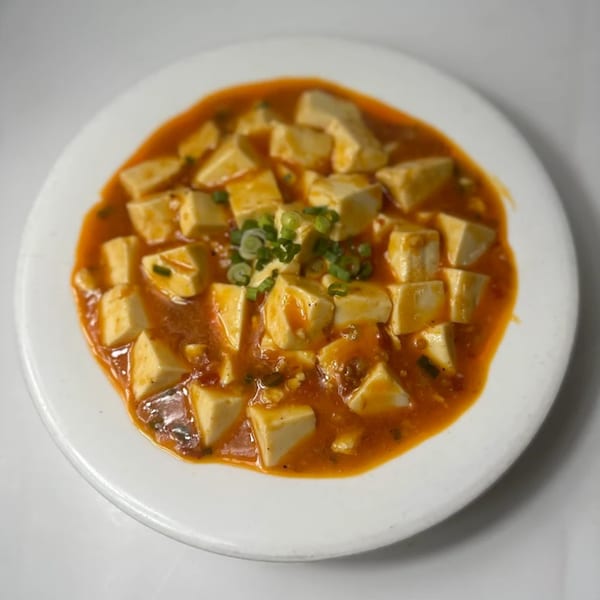 A white plate holds a dish of cubed tofu in a rich, reddish-orange sauce, garnished with green onions.